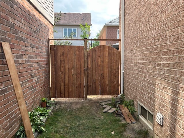 A wooden double gate made of brown planks, installed between two brick walls in a residential area with a clear sky above.
